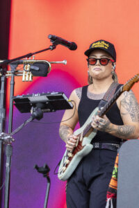 Guitarist wearing a black cap and red sunglasses plays a mint-green electric guitar on a bright orange stage backdrop.