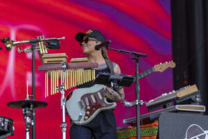 Female musician wearing a black cap and red sunglasses playing a mint-green electric guitar on a busy stage with brass instruments and keyboards around her, bright red backdrop behind.