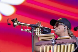 Trumpet player wearing a dark cap and sunglasses performing on a stage with bright red and pink lights in the background.