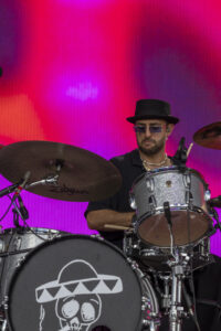 Drummer wearing a black hat and sunglasses playing a silver drum kit on a vibrant pink-purple stage backdrop with a skull logo on the bass drum.