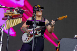 Musician on stage playing a light-blue electric guitar with a red pickguard, wearing a cap and sunglasses, surrounded by percussion and keyboard gear against a pink backdrop.