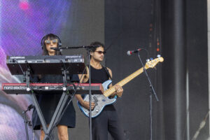 Two musicians perform on a stage: a woman at a keyboard wearing sunglasses and headphones, singing into a mic while playing a Nord Electro keyboard and a red keyboard below. A man with sunglasses plays a light blue bass guitar nearby. There is a microphone on a stand in front of them.
