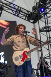 Male guitarist and singer on stage, beige shirt, singing into a microphone with hands raised.