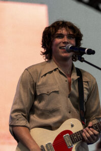 Male musician singing into a microphone while playing an electric guitar on stage, wearing a tan button-up shirt.