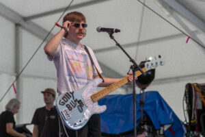 Male musician wearing sunglasses plays a light blue bass guitar at a microphone on stage under a tent.