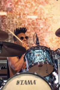 Drummer with a tall black mohawk and sunglasses, playing a turquoise Tama drum kit on a lit stage background.