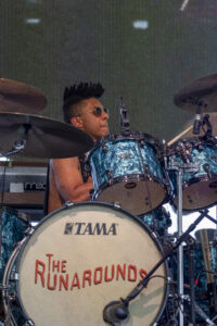 Drummer wearing sunglasses and a sleeveless shirt plays a turquoise Tama drum kit on stage, with the band's name on the bass drum head reading 'The Runarounds'.