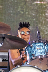 Drummer with a black mohawk and sunglasses playing a blue drum kit on stage, close-up of his face and cymbal in foreground