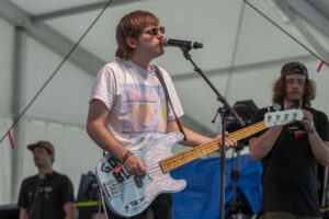 Male bassist wearing sunglasses plays a light blue bass guitar and sings into a microphone on stage.