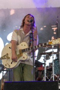Male musician singing into a microphone while playing a white electric guitar on stage.