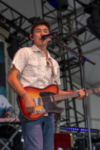 Male guitarist singing into a mic on stage, playing a sunburst electric guitar with a strap, in a concert setting.