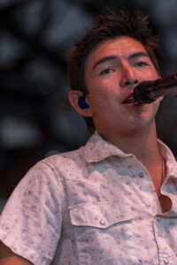 Close-up of a male singer performing on stage, singing into a microphone with a blue in-ear monitor visible in his ear.