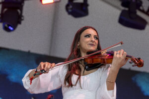 Female violinist in a white blouse plays a violin on stage under colorful lighting.