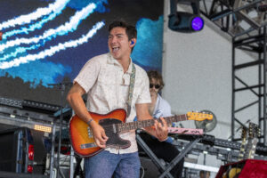 Guitarist in a light patterned shirt playing a sunburst electric guitar on a concert stage with blue LED backdrop behind him.