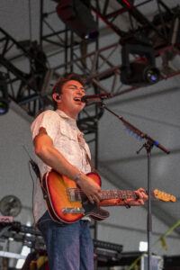 Male guitarist singing into a microphone on stage while playing a sunburst electric guitar with a strap and in-ear monitors visible.