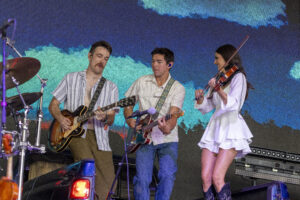 Three musicians on stage: two guitarists in the center and a violinist on the right, with a drummer partially visible on the left and colorful backdrop behind them.