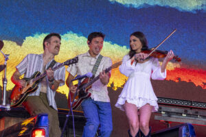 Three musicians perform on stage: two guitarists and a violinist, with a bright, multicolored backdrop behind them.