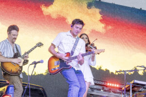 Band performing on stage: guitarist left in striped shirt, central guitarist with sunburst guitar, violinist behind, colorful backdrop behind them.