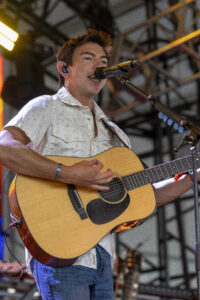 Male guitarist singing into a microphone while playing an acoustic guitar on stage in a white floral shirt.
