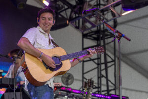 Male guitarist on stage playing an acoustic guitar and smiling, with purple stage lights behind him.