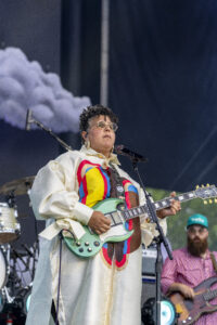 Female musician in a cream coat and round glasses plays a mint-green electric guitar on stage, singing into a microphone.