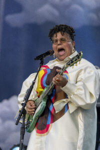 Singer with glasses passionately singing into a microphone while playing a green electric guitar on stage.