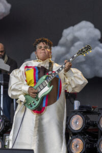 Musician on stage playing a mint-green electric guitar, wearing a cream geometric dress and round glasses.