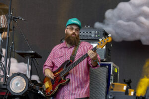 Bearded musician playing a sunburst bass guitar on stage, wearing a pink striped shirt and teal cap, with stage gear in the background.
