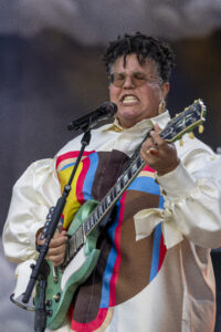 Female guitarist wearing a colorful geometric shirt plays a green electric guitar and sings into a microphone on stage.