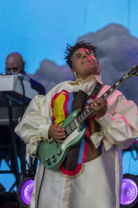 Musician playing a teal electric guitar on stage, wearing a cream shirt with rainbow stripes and round glasses against a blue backdrop.