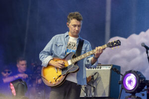 Guitarist in a denim shirt playing a sunburst electric guitar on a blue-lit stage, eyes closed in concentration.