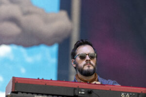 Male musician wearing sunglasses plays a red keyboard on a brightly lit stage during a live performance at a concert venue or festival.