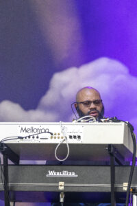 DJ with glasses and beard operates a Mellotron keyboard on a stage under purple lighting and cloud backdrop.