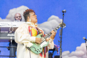 Musician in a colorful jacket playing a green electric guitar on a stage with a blue cloudy backdrop and keyboard equipment in the background.