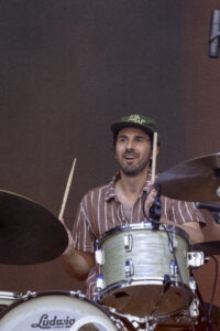Drummer wearing a green cap and striped shirt plays a Ludwig drum kit on stage.