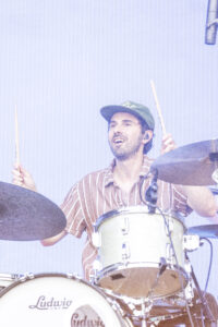 Drummer wearing a striped shirt and green cap playing a Ludwig drum kit on stage, with cymbals and a microphone nearby
