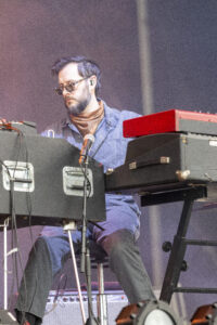Male musician wearing sunglasses and scarf playing a keyboard on stage, with red keyboard visible to the right.