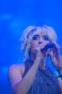 Female singer on stage, close-up of face and microphone under blue lighting, wearing a sparkly sequined top and jewelry