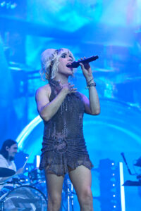 Female singer on stage wearing a sequined dress, singing into a microphone under blue stage lights.