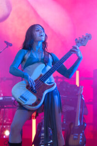 Female bassist on stage, pink and blue lighting, playing an electric bass and singing into a mic off to the side