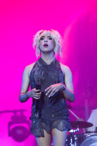 Female singer on stage, pink/purple lighting, wearing a sequined dress and holding a microphone, mid-performance.