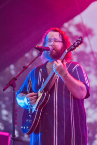 Male singer with glasses playing an electric guitar on stage, singing into a microphone under pink stage lights
