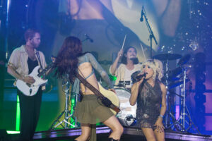 Live concert scene with a guitarist playing white electric guitar on the left and a female lead singer in a sparkling dress on the right, stage lighting in blue and purple hues behind them.