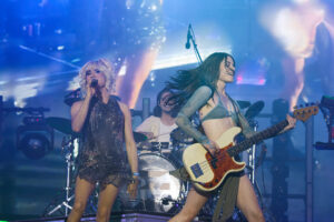 Two female musicians perform on a blue-lit stage: a singer with a microphone on the left and a bassist on the right, with a drummer in back.