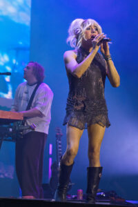 Female singer in a sparkly, sleeveless dress sings into a microphone on stage, with a keyboardist in the background under blue lights.