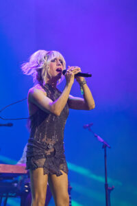 Female singer on stage wearing a glittery silver dress, holding a microphone and singing under purple-blue stage lights.