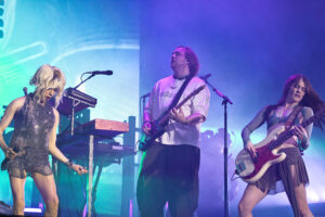 Three musicians perform on a colorful stage: a blonde female singer left, a male guitarist center, and a female bass guitarist right under blue‑purple lights.