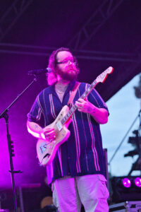 Musician with glasses and beard playing a cream-colored electric guitar on a stage bathed in purple light.