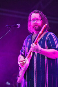 Male guitarist with glasses performing on stage under purple lighting, wearing a striped cardigan and playing a white guitar.