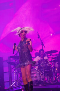 Lead singer in a glittery dress performs on a purple-lit stage, drums in the background.